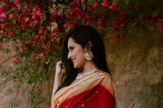 Positive young Indian lady in red traditional clothes standing near stone hill with green plants with flowers in daylight in nature